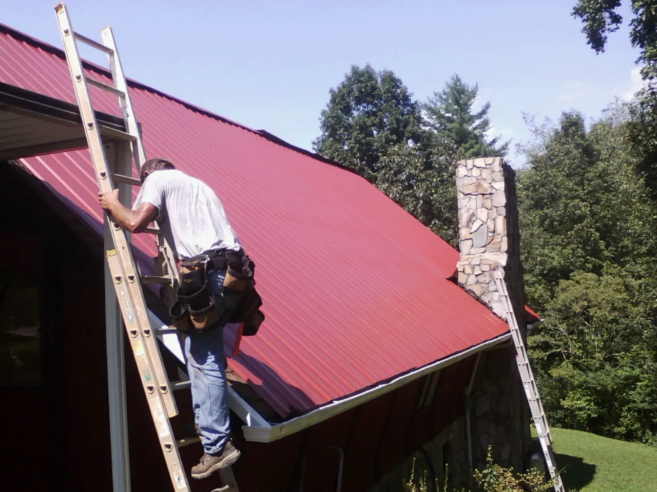 Roofer on a ladder beside a red metal roof and stone chimney — Soffit & Fascia Repair by Roofing Brothers in Mountain Home